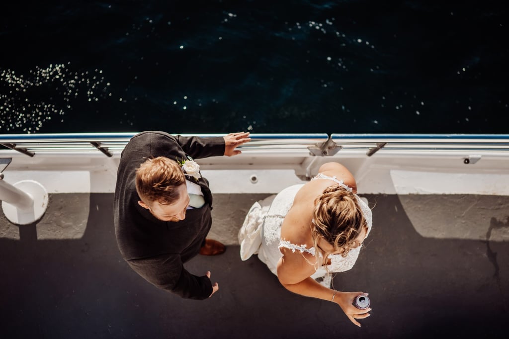 Aerial view of the bride and groom standing together on the boat deck, the sun reflecting off the deep blue ocean below.