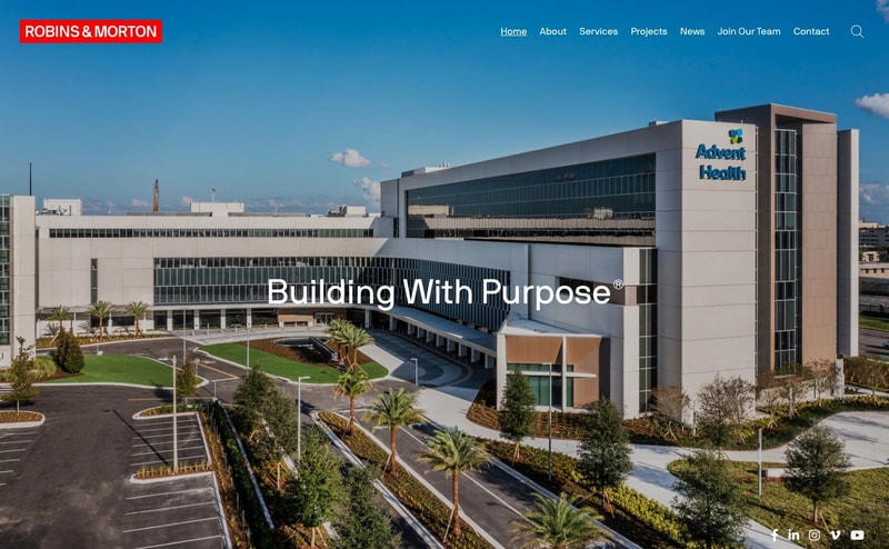 Robins & Morton homepage hero, an aerial photograph of a completed AdventHealth hospital with the single line "Building With Purpose" centered over it and a seven-item text navigation above