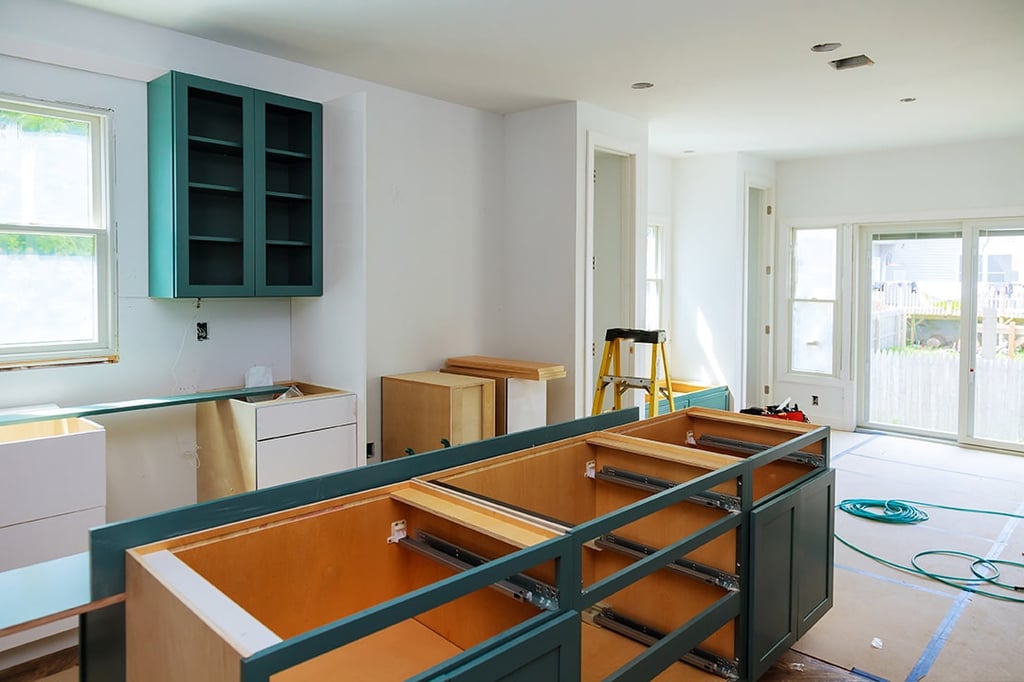A kitchen remodel underway with cabinets being installed, tools and construction materials scattered on the floor, and natural light streaming through large windows.