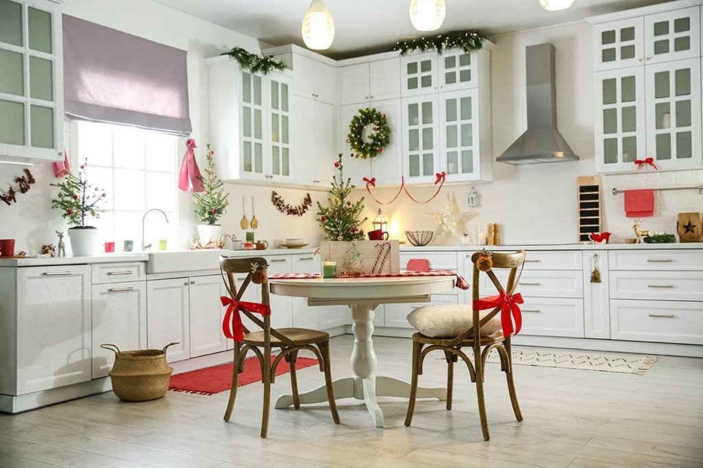 A bright, white kitchen decorated for Christmas with wreaths, ribbons, small trees, and festive table settings. Two chairs with red bows sit at a round table in the center—perfect inspiration for easy kitchen decorating and holiday ideas.