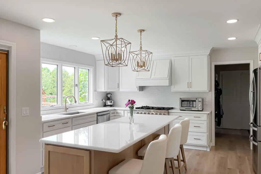 A modern kitchen by a skilled kitchen designer features white cabinets, a large island with three white chairs, gold pendant lights, stainless steel appliances, and a vase of flowers. Large windows fill the space with natural light.