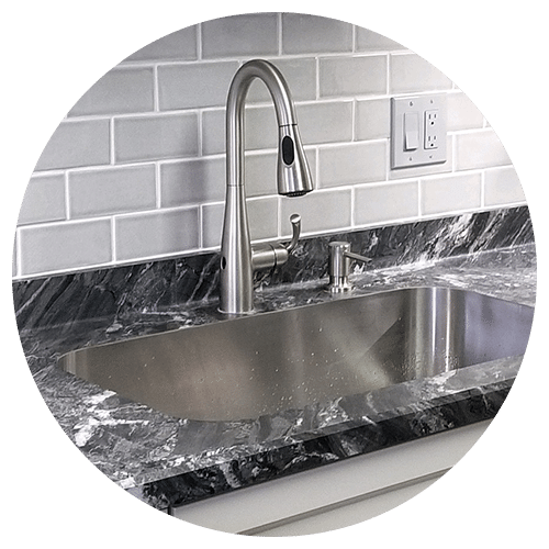 A modern stainless steel sink with a high-arc faucet is set in a dark marble countertop—an ideal centerpiece for your kitchen remodel—complete with a soap dispenser and white subway tile backsplash in the background.