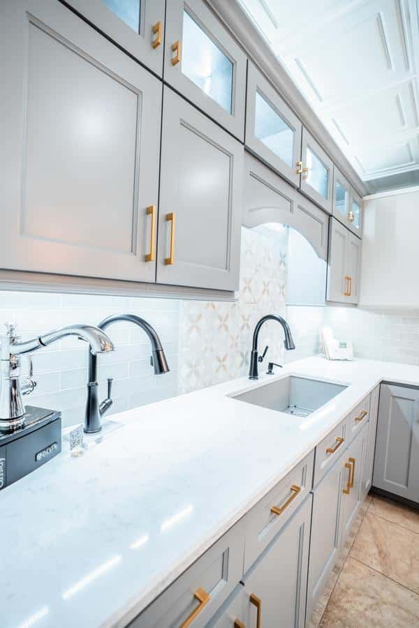 A modern kitchen with light gray cabinets, gold handles, white marble countertops, and two black faucets over a large under-mount sink—beautifully displayed at the Spencerport Showroom and brightly lit with under-cabinet lighting.
