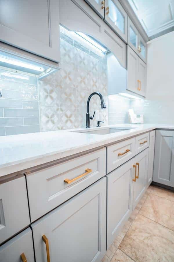 Modern kitchen with white cabinets, gold handles, a marble countertop, black faucet, and decorative tile backsplash—beautifully displayed in our Spencerport Showroom and brightly lit by under-cabinet lights.