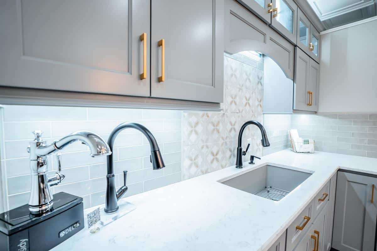 Modern kitchen with light gray cabinets, gold handles, a white marble countertop, two black faucets, and a stainless steel sink. The space is brightly lit, showcasing the light tile backsplash with geometric patterns in our Spencerport Showroom.