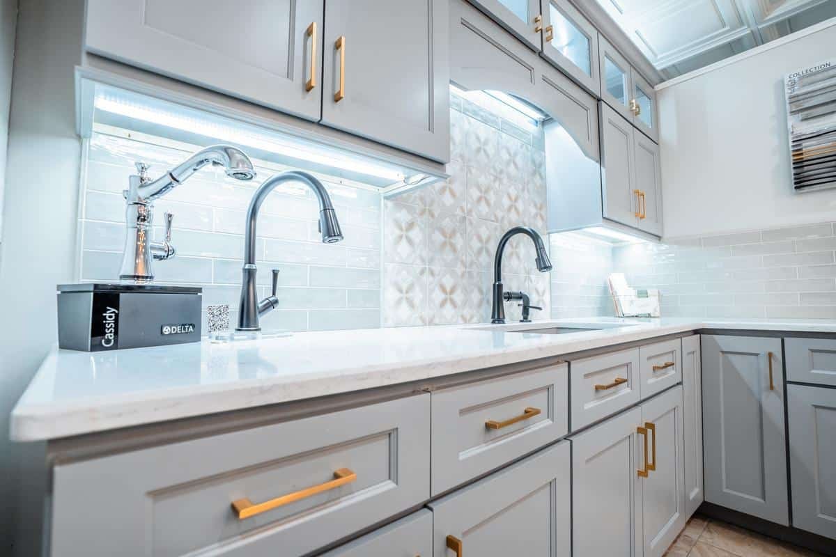Modern kitchen with light gray cabinets, gold handles, white marble countertop, under-cabinet lighting, two black faucets, tiled backsplash, and a Delta faucet box on the counter at the Spencerport Showroom.