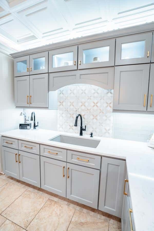 Modern kitchen in our Spencerport Showroom with gray cabinets, gold handles, white countertops, black faucet, patterned tile backsplash, and under-cabinet lighting. Upper cabinets feature glass inserts displaying white dishes.