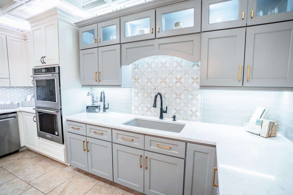 Modern kitchen with light gray cabinets, gold handles, white countertops, double wall ovens, a large sink, black faucet, under-cabinet lighting, and a patterned tile backsplash—see it in person at our Spencerport Showroom.