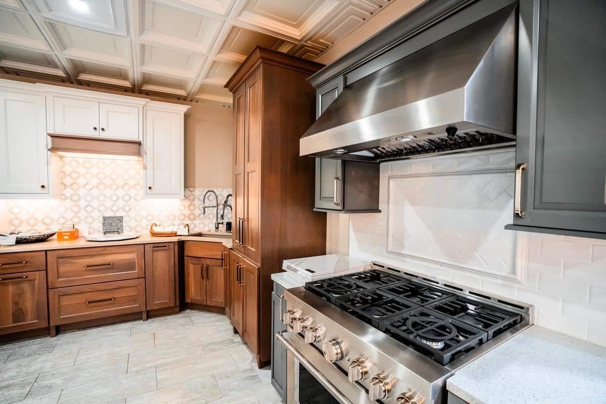 Modern kitchen with stainless steel gas stove and range hood, brown wooden cabinets, white backsplash tiles, and light countertops. Geometric tile backsplash and under-cabinet lighting complete this stylish Spencerport Showroom display.