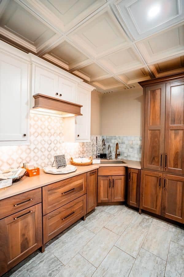 Modern kitchen with wood cabinets, white upper cupboards, a patterned tile backsplash, and light-colored tile flooring. Perfect for a showroom in Spencerport, the countertops feature a tray, decor items, and a corner sink with bronze faucet.