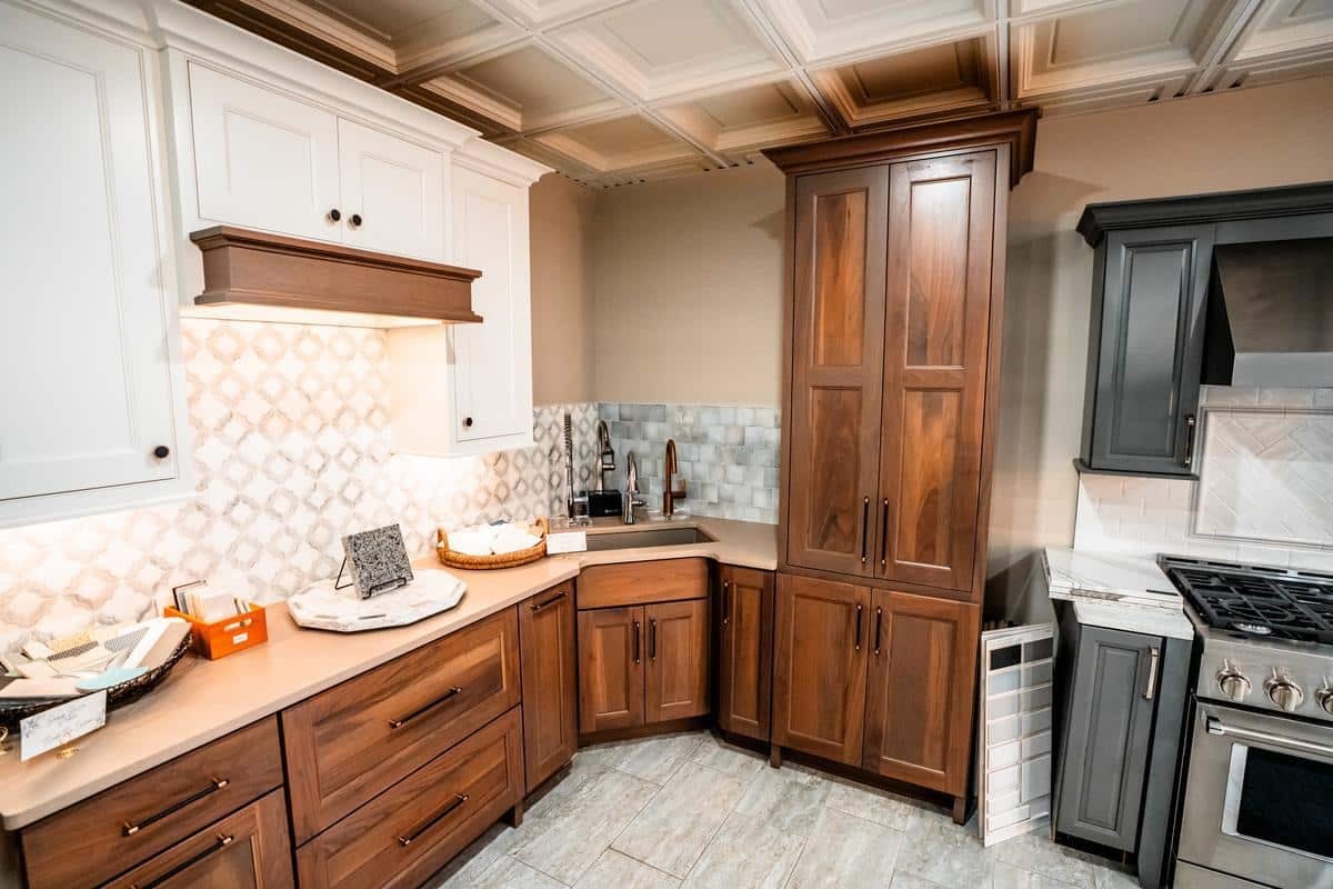 A modern kitchen features wood cabinets, a white tile backsplash with patterned accents, stainless steel appliances, and light countertops. The well-lit space is organized with various cooking essentials on the counters—just like the Spencerport Showroom.