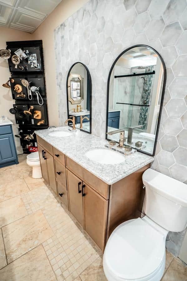 A modern bathroom with a marble countertop, double sinks, two arched mirrors, a wooden vanity, a white toilet, and hexagon wall tiles. Fixtures and accessories from the Spencerport Showroom are displayed on a wall rack in the background.
