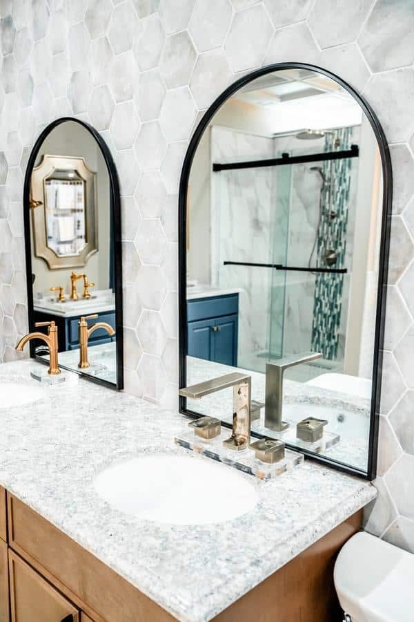 Modern bathroom with two sinks, granite countertop, arched mirrors, gold and silver faucets, and hexagonal wall tiles—plus a glimpse of the shower and blue cabinetry reflected in the mirrors at the Spencerport Showroom.