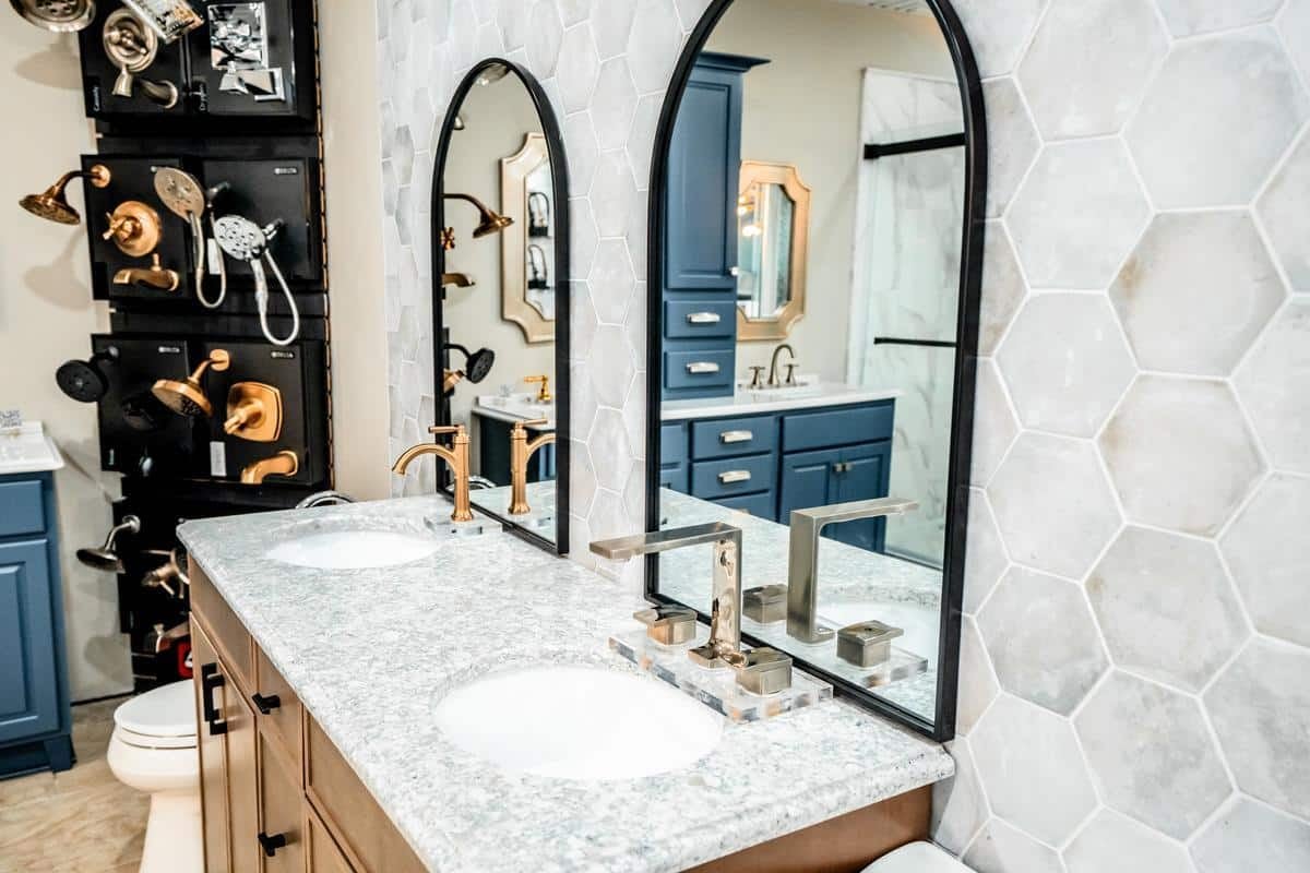 Modern bathroom with a double sink vanity, marble countertop, blue cabinets, and hexagonal light gray wall tiles. Fixtures are displayed on the left wall in our Spencerport Showroom, featuring gold and silver faucets and two arched mirrors.