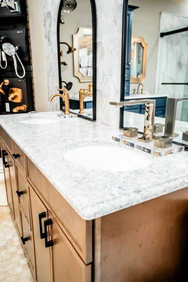 Modern Spencerport showroom display featuring a double vanity with a speckled white countertop, two sinks, gold and silver faucets, wooden cabinets, and decorative mirrors against a tile backsplash.