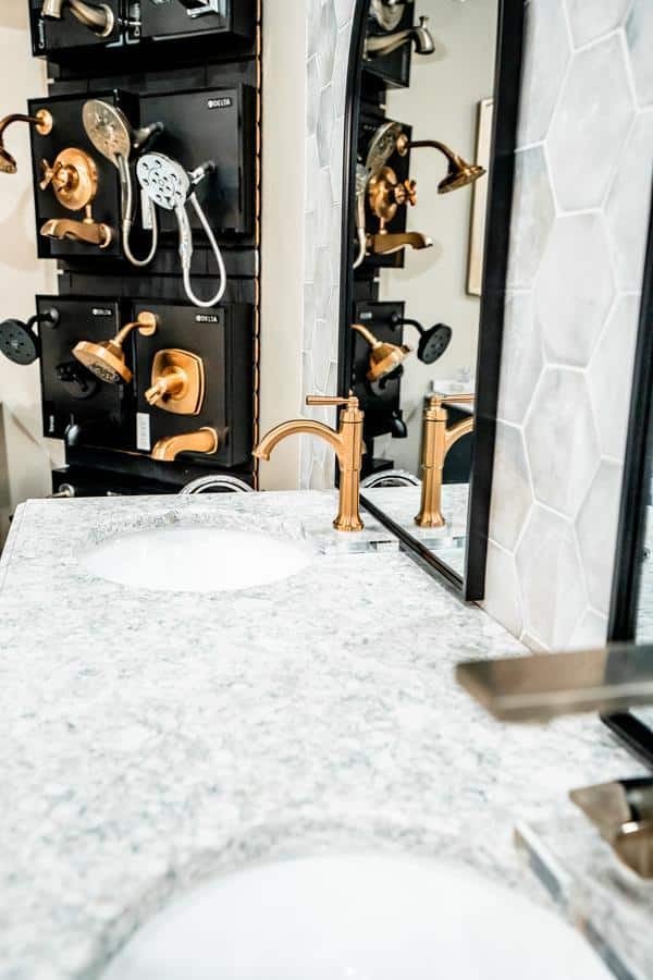A bathroom countertop with a white sink, gold faucet, and a decorative hexagon-patterned tile backsplash. In the mirror’s reflection, showroom displays of various shower fixtures are visible in Spencerport.