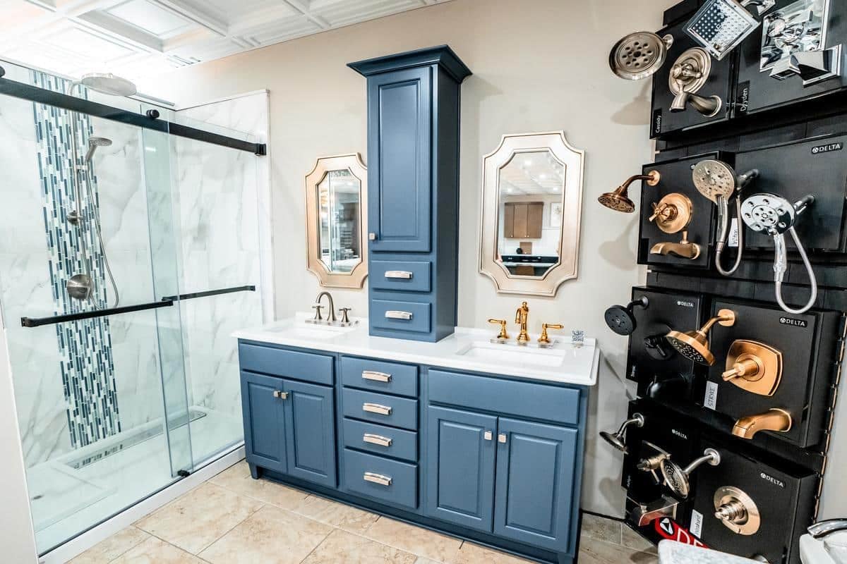 A modern Spencerport Showroom displays a blue double-sink vanity with gold faucets, two mirrors, and a tall central cabinet. On the right, various showerheads and faucets are mounted on a display wall.