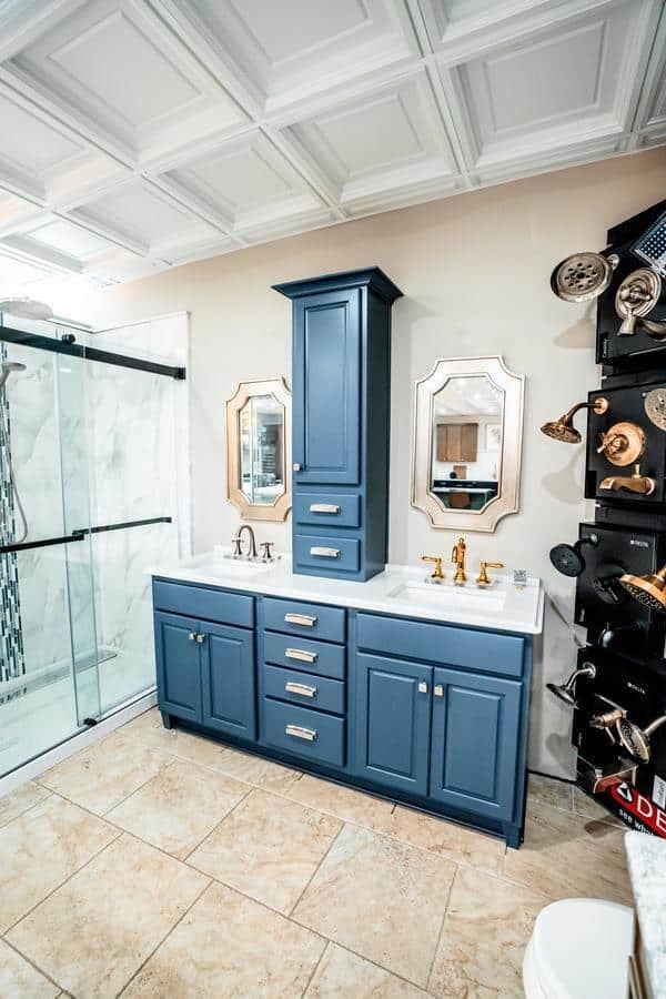 Modern bathroom with a blue double-sink vanity, two mirrors, gold faucets, beige tile floor, and a glass shower. Explore wall-mounted showerheads on the right at the Spencerport Showroom.