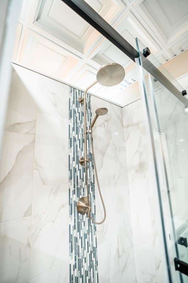 A modern shower with a rainfall showerhead and handheld sprayer, set against white marble tiles with a vertical strip of blue and gray mosaic accents. Featured in our Spencerport Showroom, with a glass door and paneled ceiling also visible.