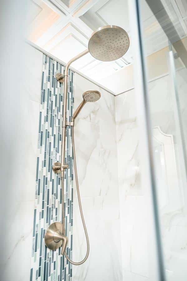 Modern shower with a large round overhead showerhead and a detachable handheld shower on a vertical rail, featured in our Spencerport showroom, set against white marble tiles with a vertical strip of blue and gray mosaic tiles.
