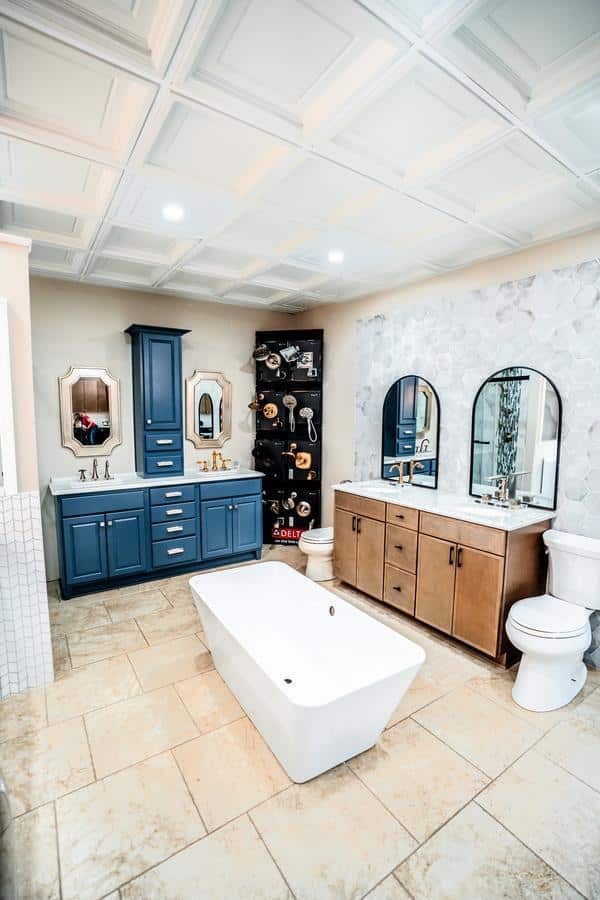 The Spencerport Showroom features a modern bathroom display with a freestanding bathtub, blue and brown vanities with matching mirrors, two toilets, marble tile flooring, and a decorative wall of faucets and showerheads.