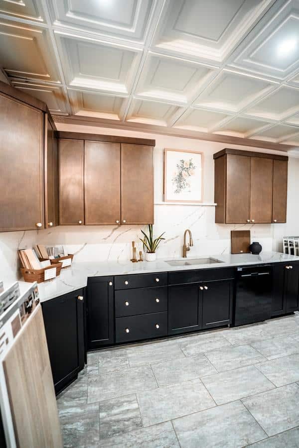 Modern kitchen with brown upper cabinets, black lower cabinets, white marble backsplash, gold fixtures, and a plant by the sink. Art on the wall and gray tile floors complete this stylish look from our Spencerport Showroom.