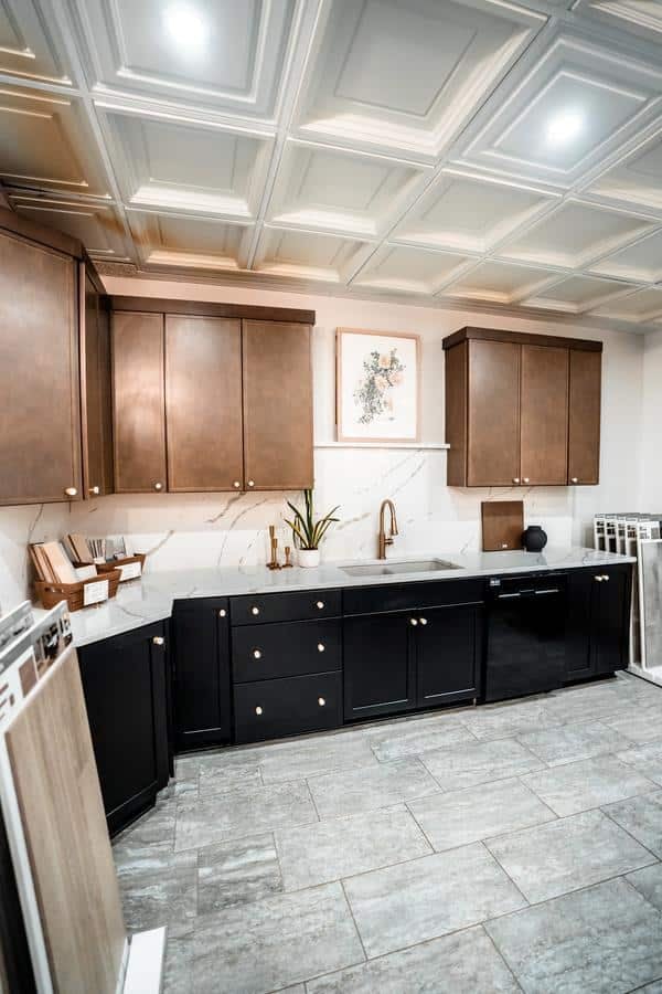 A modern kitchen at the Spencerport Showroom features brown upper cabinets, black lower cabinets, a gray tile floor, and a white marble backsplash. Gold hardware accents the space, with a plant and artwork above the sink and samples displayed nearby.