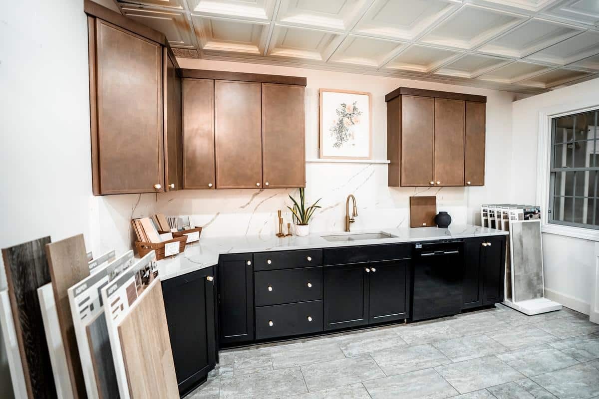 Modern Spencerport showroom featuring brown upper cabinets, black lower cabinets, marble countertops, a gold faucet, and sample boards of flooring and tile on each side. A plant and framed art are displayed above the sink.