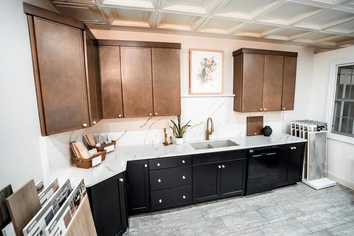 Modern kitchen in the Spencerport Showroom with upper brown cabinets, lower black cabinets, a white marble countertop, stainless steel sink, and sample tiles on the floor. A small plant and wall art add stylish touches to the space.