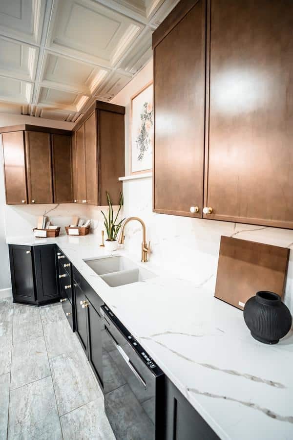 Modern kitchen with brown upper cabinets, black lower cabinets, white marble countertops, brass faucet, green plant, framed artwork, and decor on the counter—featured in the Spencerport Showroom—set on light gray tiled flooring.