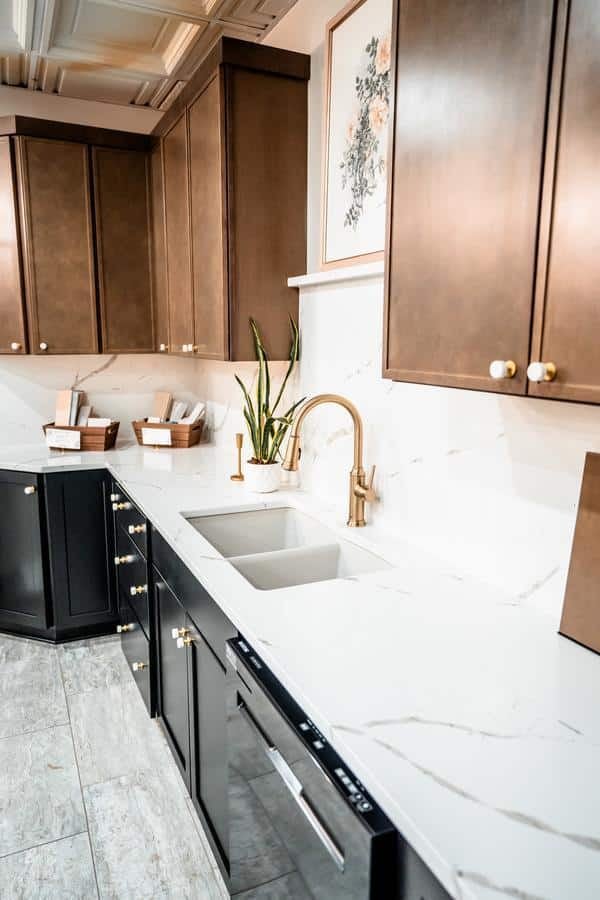 Modern kitchen with brown upper cabinets, black lower cabinets, gold hardware, a marble countertop and backsplash, a built-in sink with a gold faucet, and framed floral artwork above the counter—discover inspiration at our Spencerport Showroom.