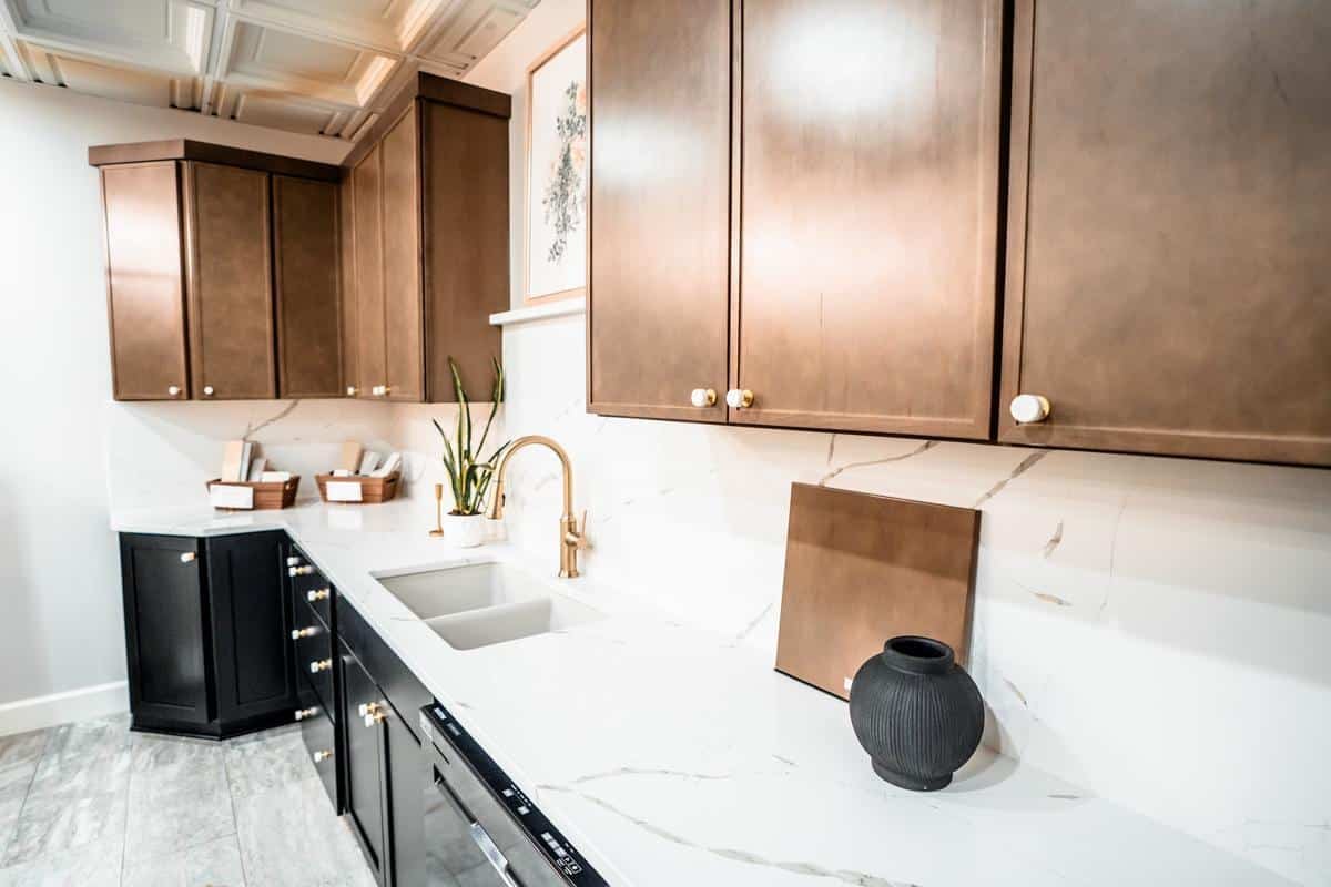 Modern kitchen showroom in Spencerport with brown upper cabinets, black lower cabinets, white marble countertops and backsplash, gold faucet, a black vase, two baskets, plants, and framed art on the wall.