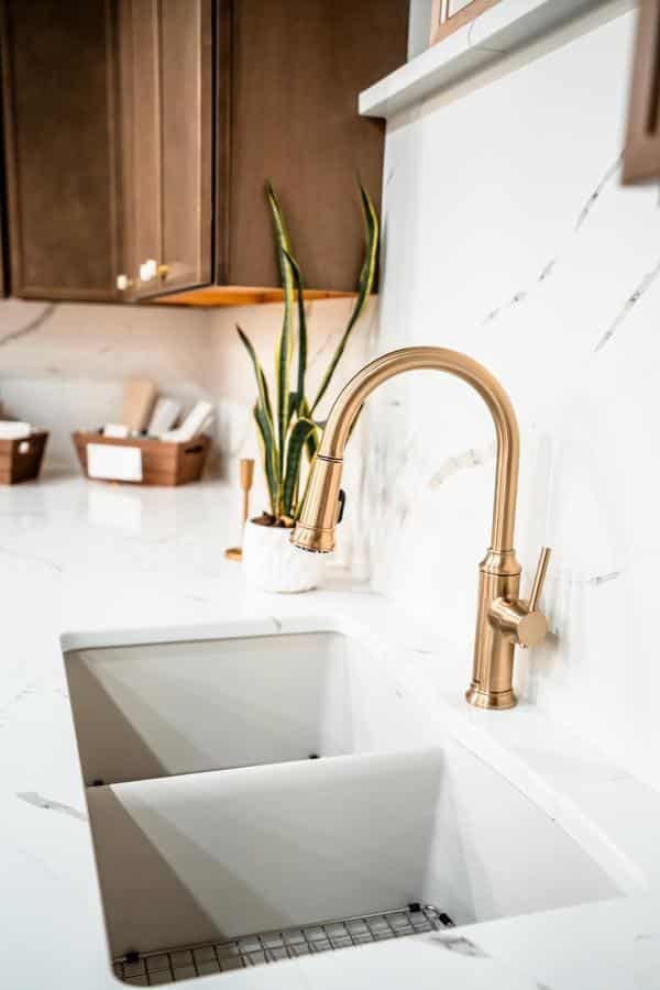 A modern kitchen sink with a gold faucet, white marble countertop, and a potted snake plant. Wooden cabinets and organized baskets are visible in the background, showcasing inspiration from the Spencerport Showroom.