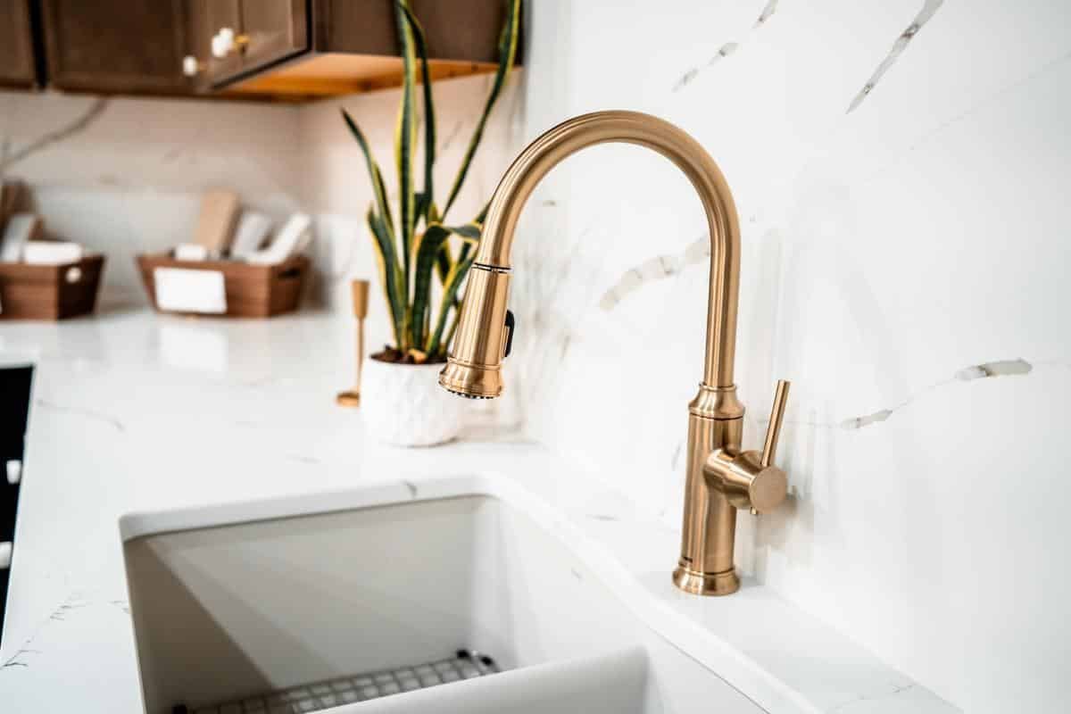 A modern kitchen sink with a gold faucet, white marble countertop, potted snake plant, and brown cabinets in the background—visit our Spencerport Showroom to explore similar design inspirations.