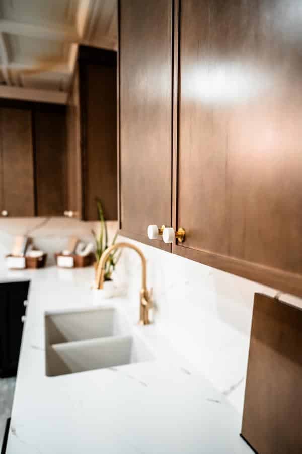 A modern kitchen with dark wooden cabinets, a white marble countertop, and gold faucet—see similar designs at the Spencerport Showroom, where small potted plants and containers add charm along the backsplash.