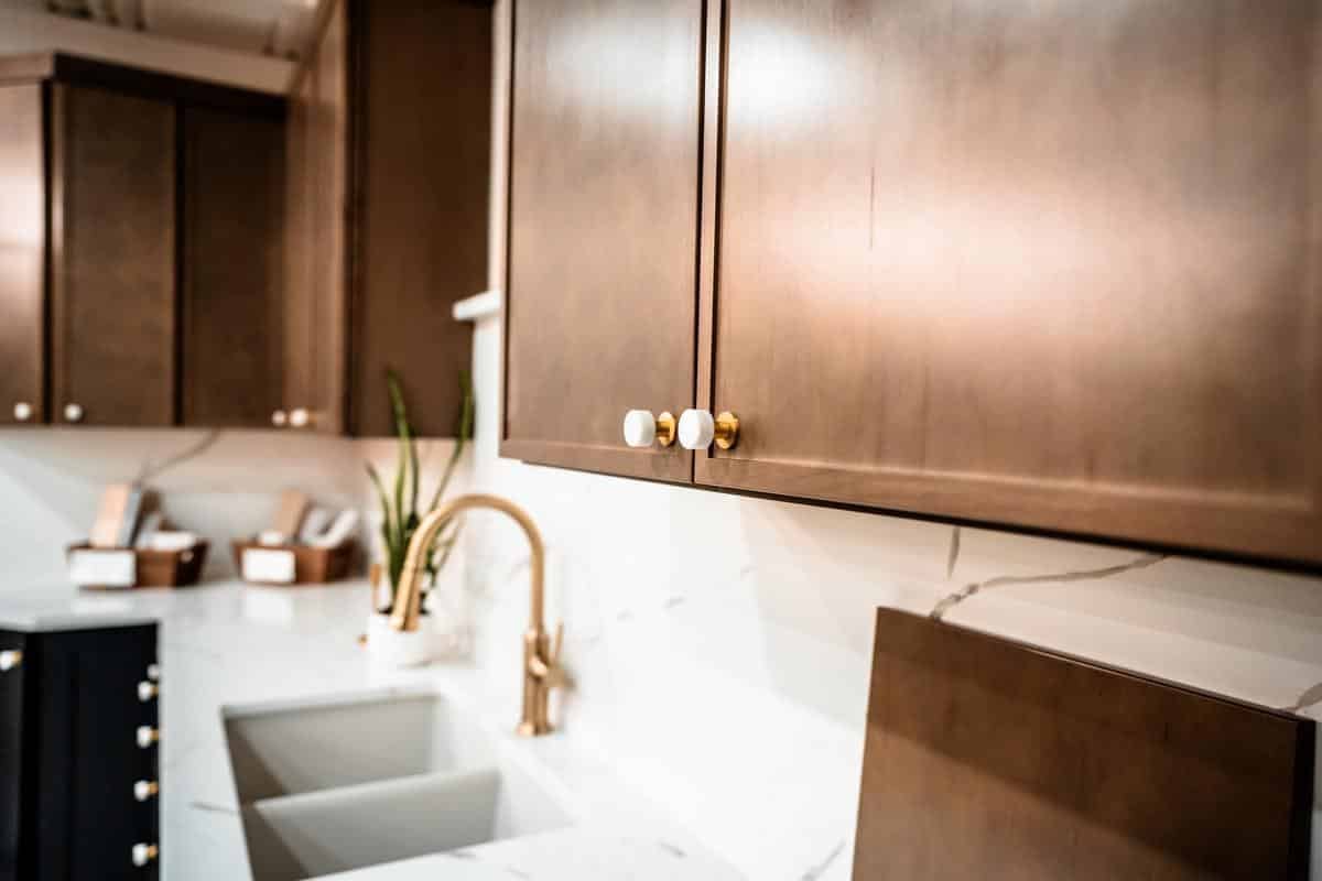 Modern kitchen with light brown cabinets, white marble countertops, a gold faucet, and double sink. A plant sits near the sink, while boxes line the counter in the background—all showcased at our Spencerport Showroom.