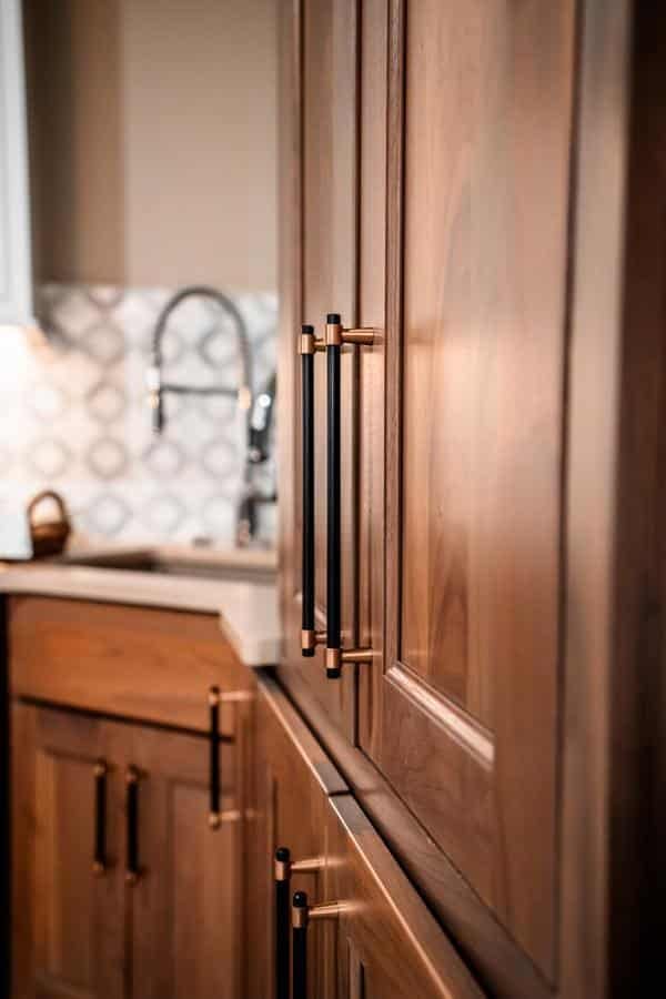 Close-up of wooden kitchen cabinets with modern black and gold handles, showcased in the Spencerport Showroom, with a sink and patterned backsplash visible in the background.