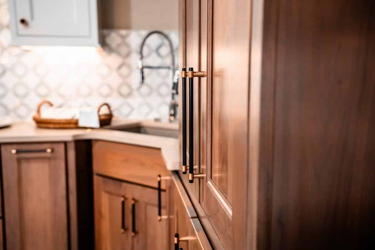 Close-up of modern wooden kitchen cabinets with gold handles at the Spencerport Showroom. In the background, a countertop, woven basket, patterned backsplash, and a sink with a gooseneck faucet complete the look.