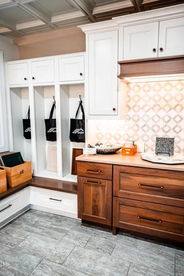 A modern kitchen, inspired by the Spencerport Showroom, features white upper cabinets, wooden lower cabinets, a patterned tile backsplash, and a built-in bench with storage cubbies and hooks holding black tote bags. The floor is light gray tile.
