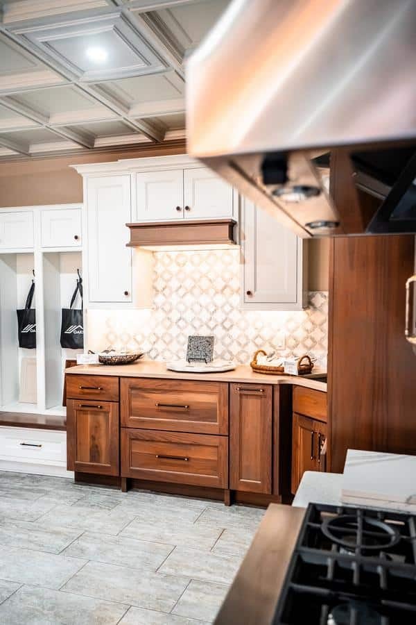 A modern kitchen in the Spencerport Showroom features wooden lower cabinets, white upper cabinets, a geometric tile backsplash, built-in lighting, and a stainless steel stove in the foreground, with a bench and storage area to the left.
