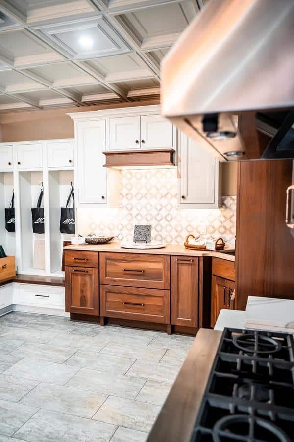 A modern kitchen with wood and white cabinets, a patterned backsplash, a gas stove in the foreground, and a built-in bench with coat hooks and storage on the left side—featured in our Spencerport Showroom.