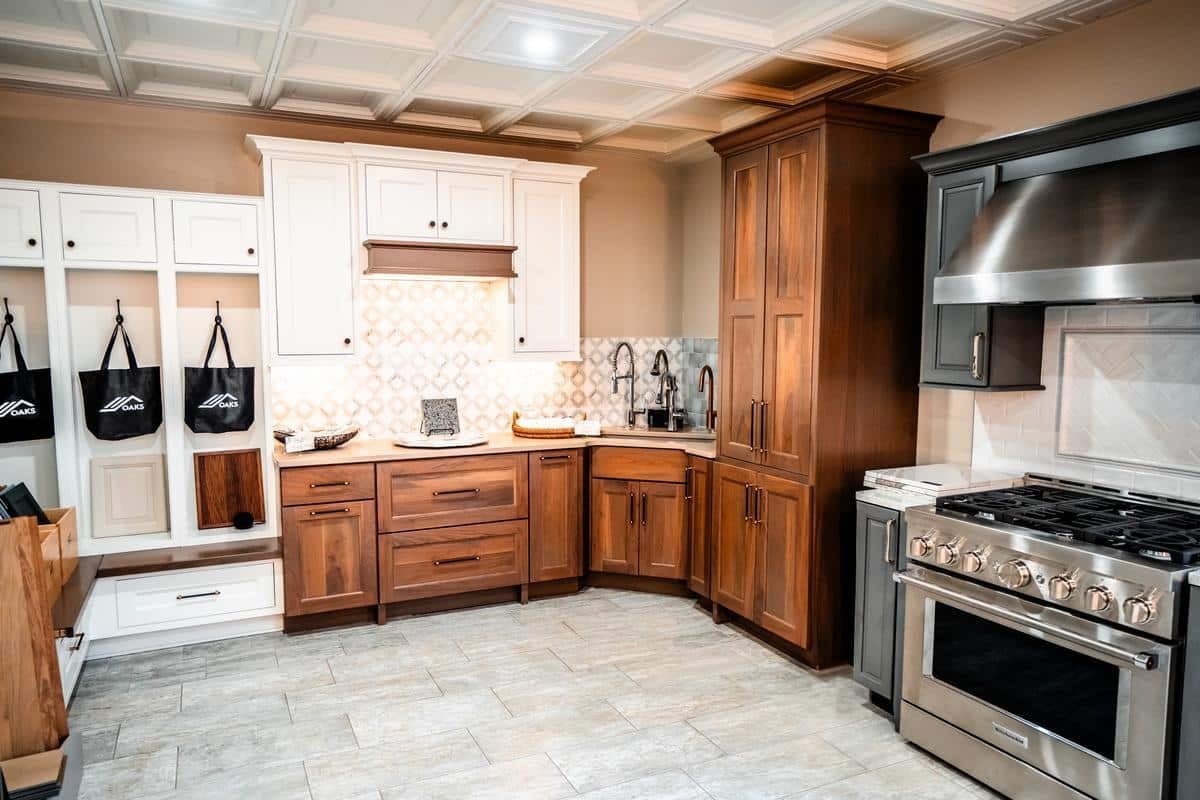 Modern kitchen showroom in Spencerport featuring wooden cabinets, stainless steel stove and range hood, patterned tile backsplash, and a white mudroom area with black hanging bags. Light-colored tile flooring runs throughout.