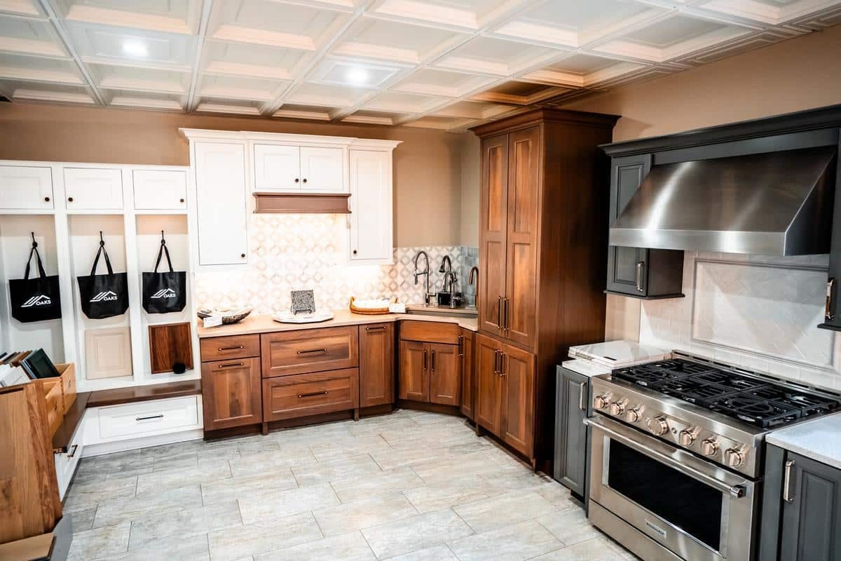 Modern kitchen showroom in Spencerport featuring white and dark wood cabinets, stainless steel appliances, a large oven with hood, tiled backsplash, and hooks holding black aprons on the left wall. The floor is light-colored tile.
