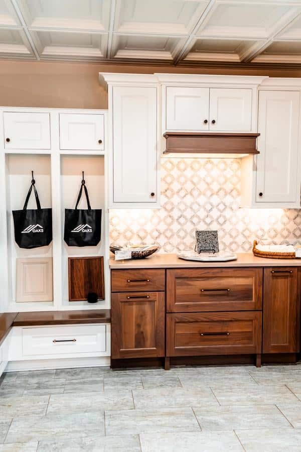A kitchen inspired by the Spencerport Showroom features white upper cabinets, wood lower cabinets, a patterned tile backsplash, and light-colored tile flooring. Hooks on the side wall hold two black bags above a built-in bench with storage.