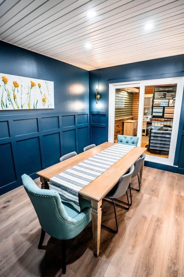 A modern meeting room in the Spencerport Showroom features dark blue paneled walls, a wooden table with a striped runner, four gray chairs, two teal armchairs, wood flooring, a floral painting, and bright overhead lighting.
