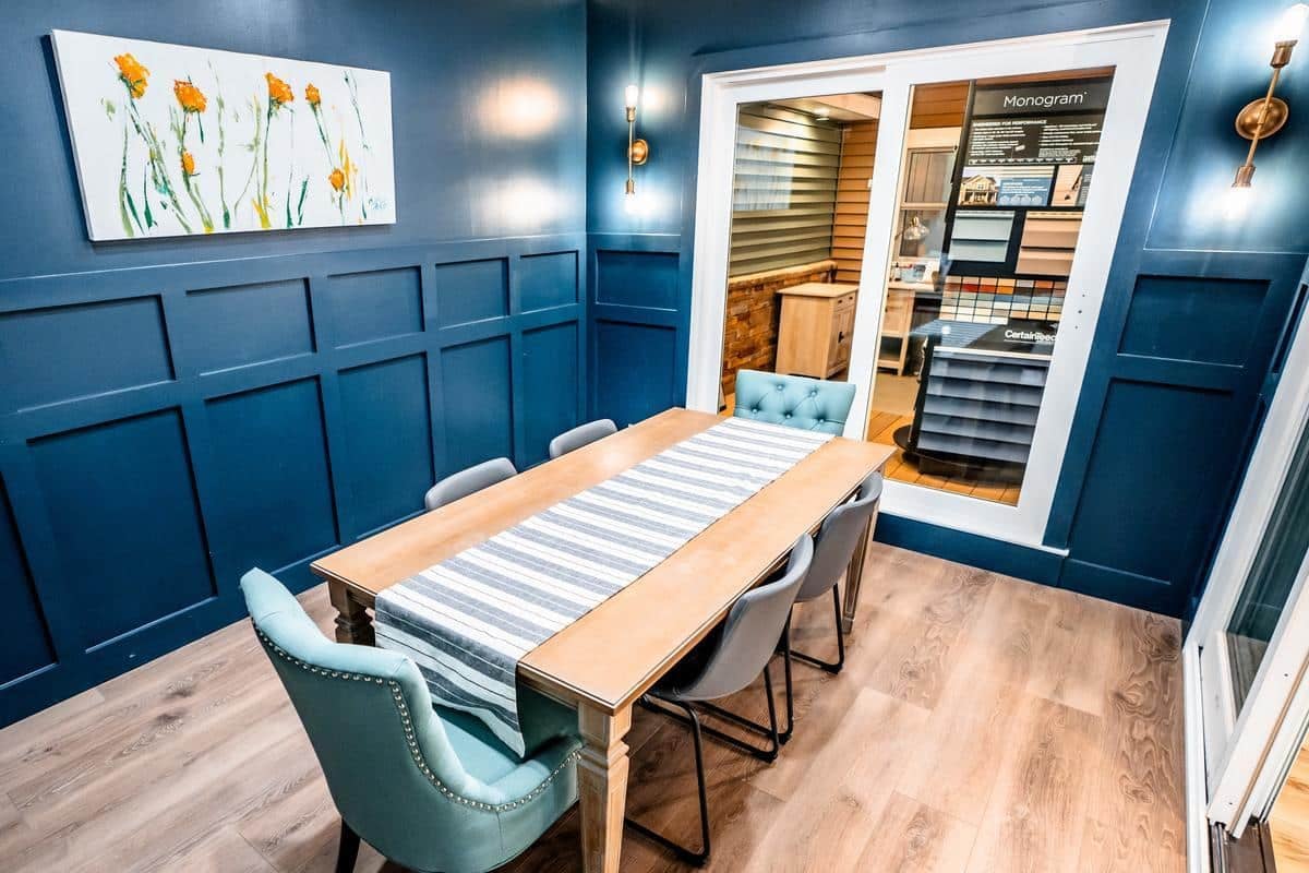 A modern dining room with blue paneled walls, a wooden table with a striped runner, four gray chairs, and a blue upholstered chair. Wall sconces and a floral painting add charm. Sliding glass doors reveal the Spencerport Showroom beyond.