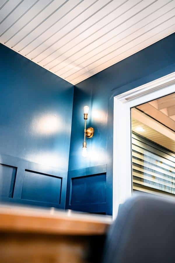 A modern interior corner in Spencerport with blue paneled walls, a white paneled ceiling, a brass wall sconce with two exposed bulbs, and a window reflecting light from another room—perfect for showroom inspiration.