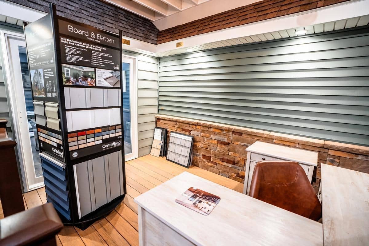 The Spencerport Showroom displays home siding options, including board and batten styles, stone veneer, and paint samples. Two white desks, a brown chair, and sample boards are arranged on the wall and floor.