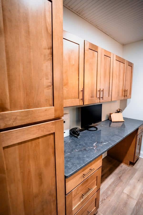 A home office or workspace with light wood cabinets, drawers, and a dark countertop, inspired by the Spencerport Showroom. A computer monitor, keyboard, and cutting board sit on the counter above the light wood floor.