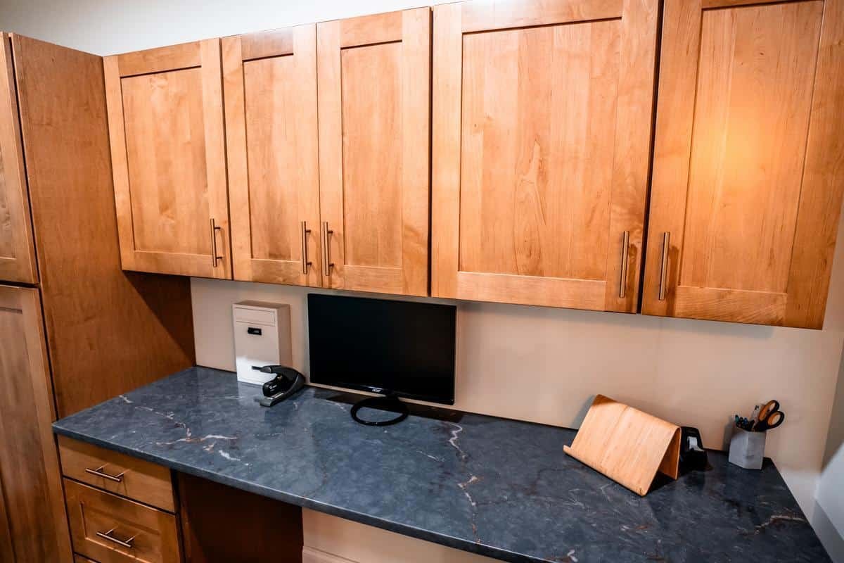 A modern workspace in the Spencerport Showroom showcases light wood cabinets above a dark marble countertop, featuring a closed laptop, monitor, phone, pen holder, and a small wooden stand.
